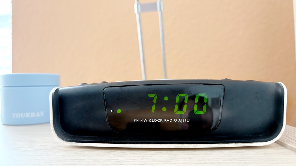 Ein digitaler Wecker zeigt 7:00 Uhr. Daneben steht eine hellblaue Dose mit der Aufschrift YOURDAY auf einem Holz-Nachttisch – Symbol für den Start einer strukturierten Morgenroutine.
